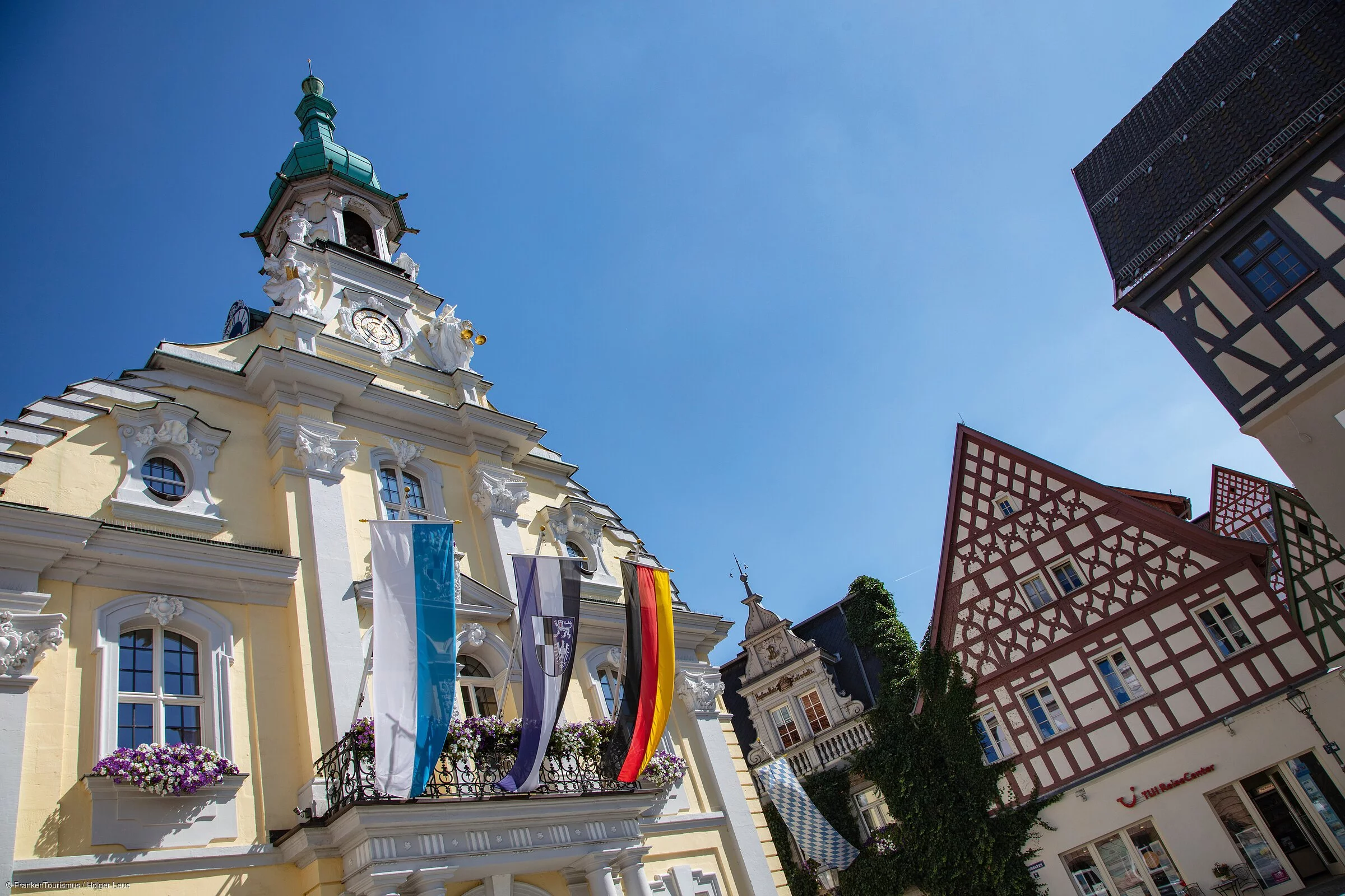 Ein prächtiges, historisches Gebäude in gelblicher Farbe, geschmückt mit Fahnen, daneben Fachwerkbauten