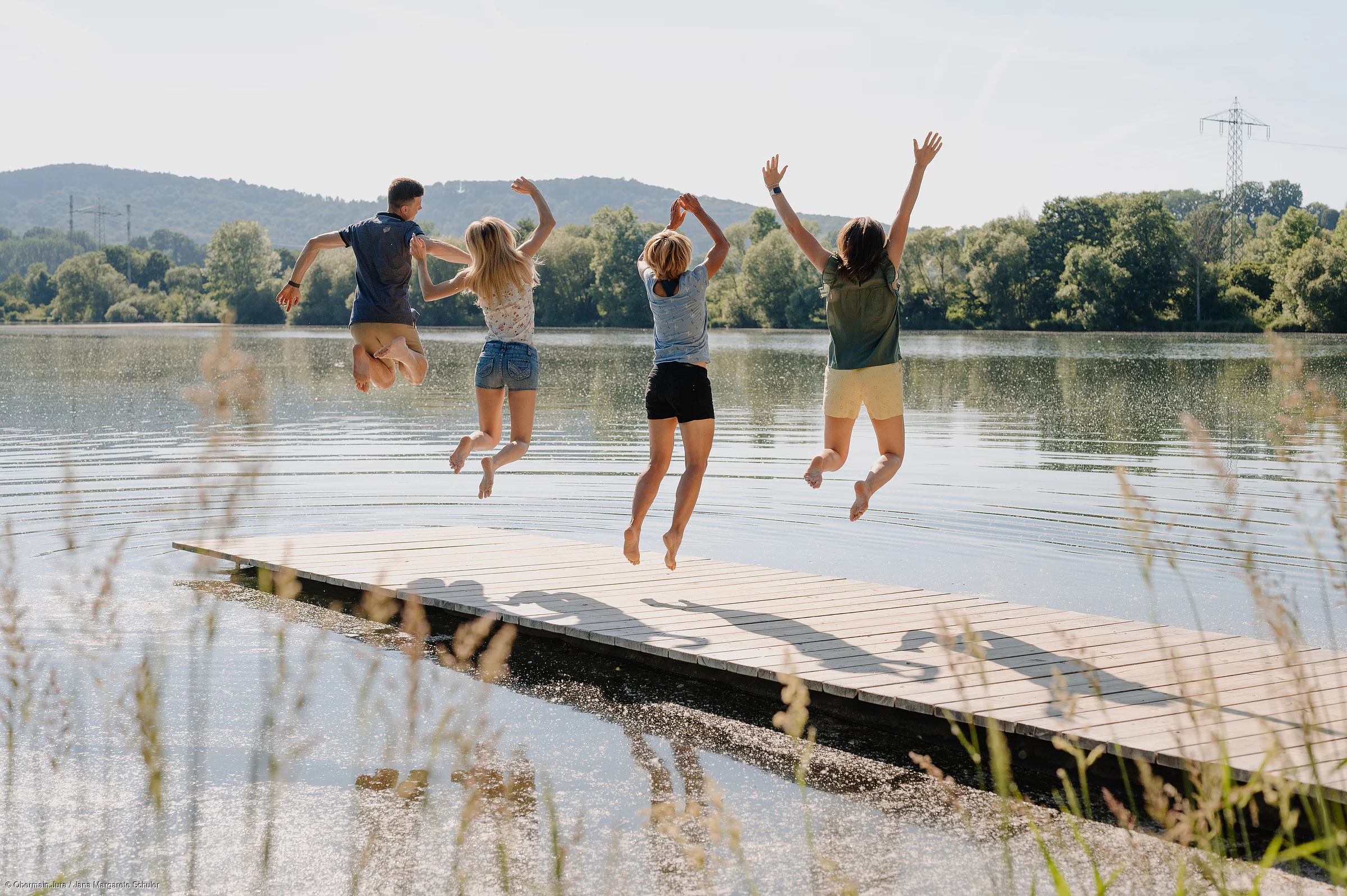 Vier junge Menschen springen auf einem Steg in die Luft, im Hintergrund ein See