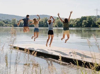 Vier junge Menschen springen auf einem Steg in die Luft, im Hintergrund ein See