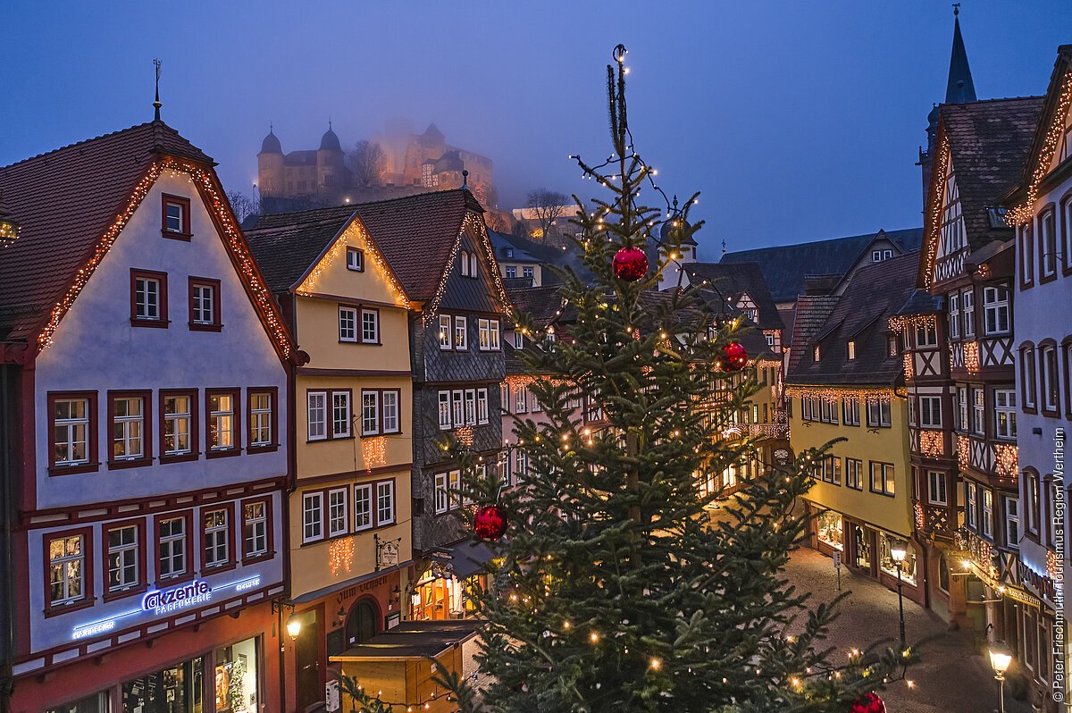 Beleuchtete Fachwerkhäuser und ein geschmückter Weihnachtsbaum in einer abendlichen Stadtansicht.