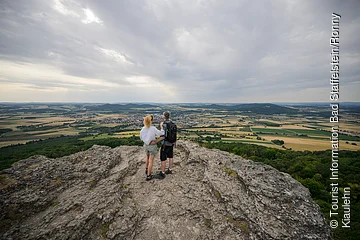 Zwei Personen stehen auf einem Felsen und blicken auf eine weite Landschaft mit Feldern und einem Dorf.