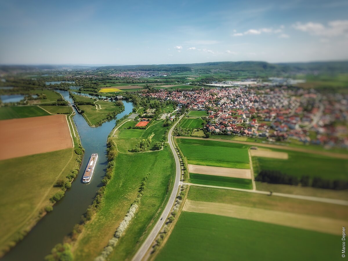 Luftaufnahme einer Flusslandschaft mit einem Frachtschiff, umgeben von Feldern und einer Siedlung im Hintergrund.