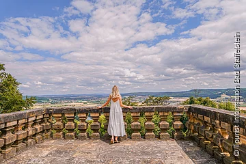 Person in weißem Kleid steht auf einer Steinplattform und blickt auf eine weite Landschaft mit Hügeln und Wolken.