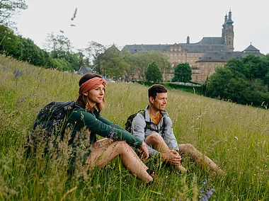Zwei Wandernde machen Rast auf einer Wiese, im Hintergrund eine Klosteranlage mit Türmen