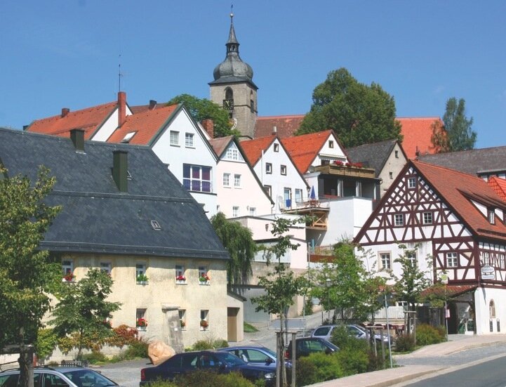 Blick auf ein Dorf mit Fachwerkhäusern, Bäumen, parkenden Autos und Kirchturm bei blauem Himmel.