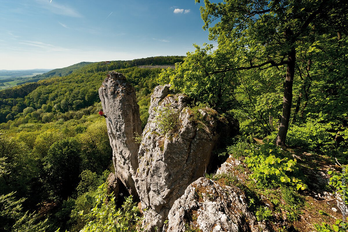 Felsformation im Wald mit Kletterer an einer steilen Felswand bei klarem Himmel.