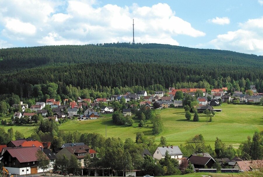 Bischofsgrün (Fichtelgebirge) Dorf mit Häusern am Waldrand vor bewaldetem Hügel und Sendemast unter blauem Himmel mit Wolken