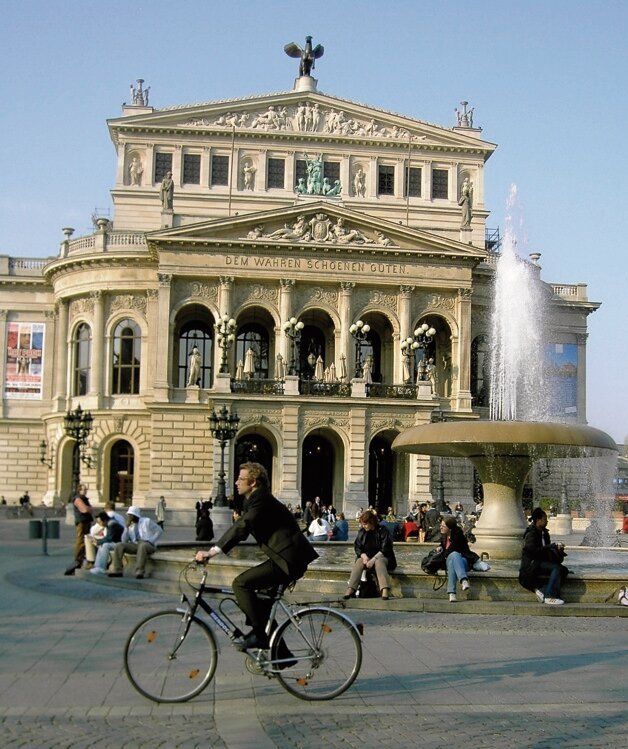 Fahrradfahrer vor historischem Gebäude mit Brunnen, Menschen sitzen am Brunnenrand, sonniger Tag.