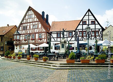 Marktplatz mit Fachwerkhäusern, Sitzgelegenheiten, Sonnenschirmen und einer Statue in der Mitte