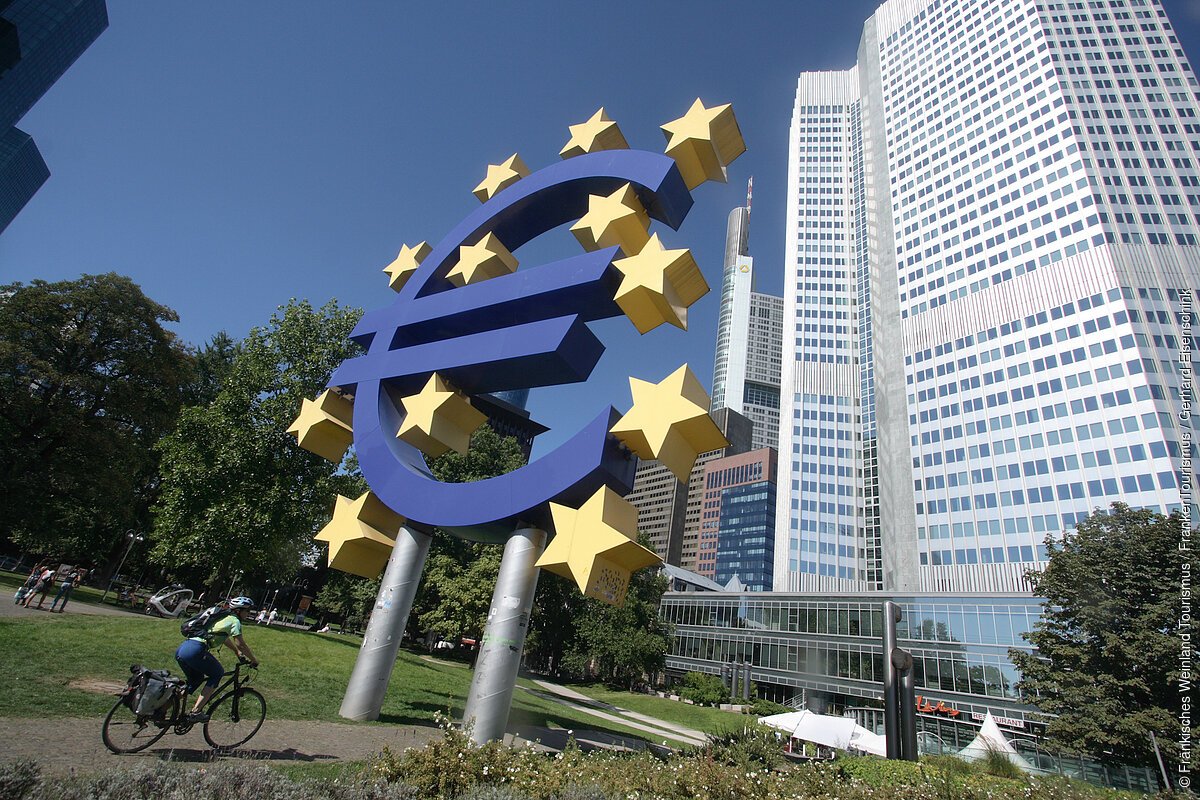 Große Euro-Skulptur mit gelben Sternen vor Hochhäusern und Radfahrer auf einem Weg im Park.