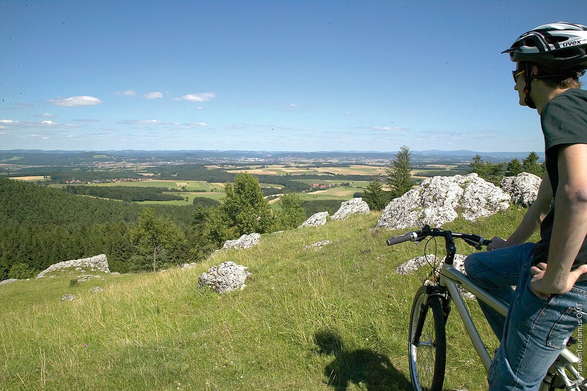 Person mit Fahrrad und Helm blickt auf grüne Wiesen, Felsen und weite Landschaft unter blauem Himmel