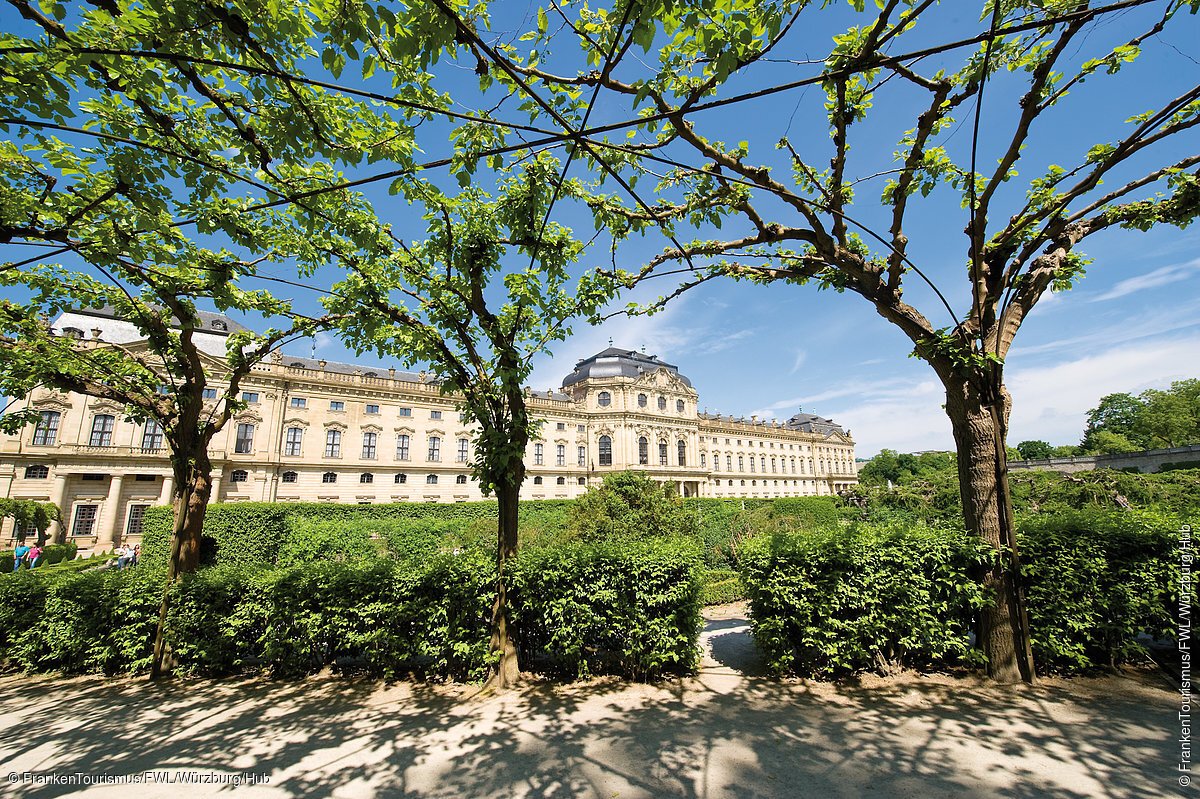 Residenz und Hofgarten (Würzburg, Fränkisches Weinland) Blick durch grüne Baumreihen auf ein historisches Schlossgebäude bei blauem Himmel.