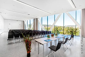 Konferenzraum mit Stuhlreihen, Tischen mit Unterlagen und großen Fenstern mit Stadtblick.